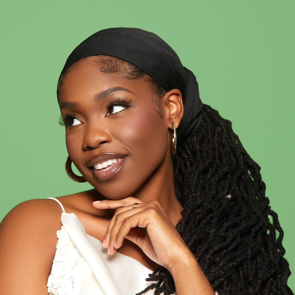Woman with dark skin wearing a black satin wrap scarf tied over long dreadlocks, gold hoop earrings, and a white sleeveless top against a green background.