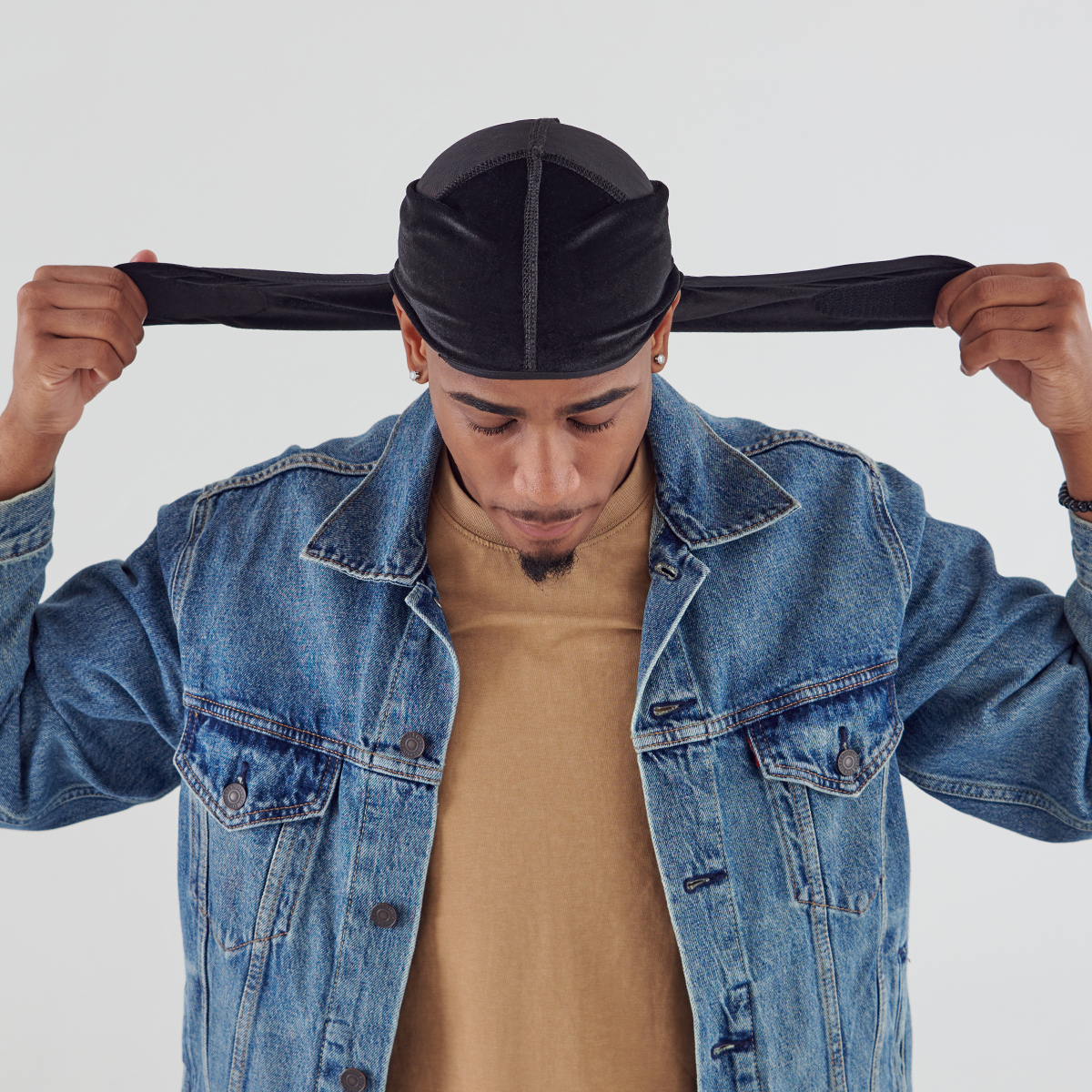 Man wearing a black velvet durag with extended straps, pulling the straps outward while looking down, dressed in a tan shirt and denim jacket.