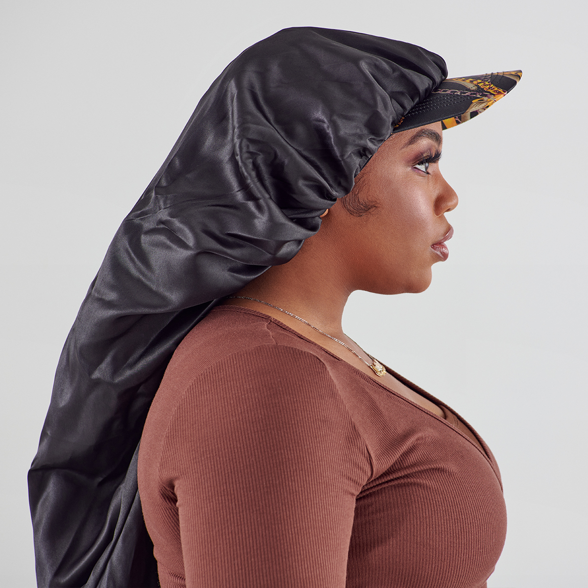 Profile view of a woman wearing a black silky satin bonnet cap with a visor featuring a patterned design, styled over her hair, against a plain background.