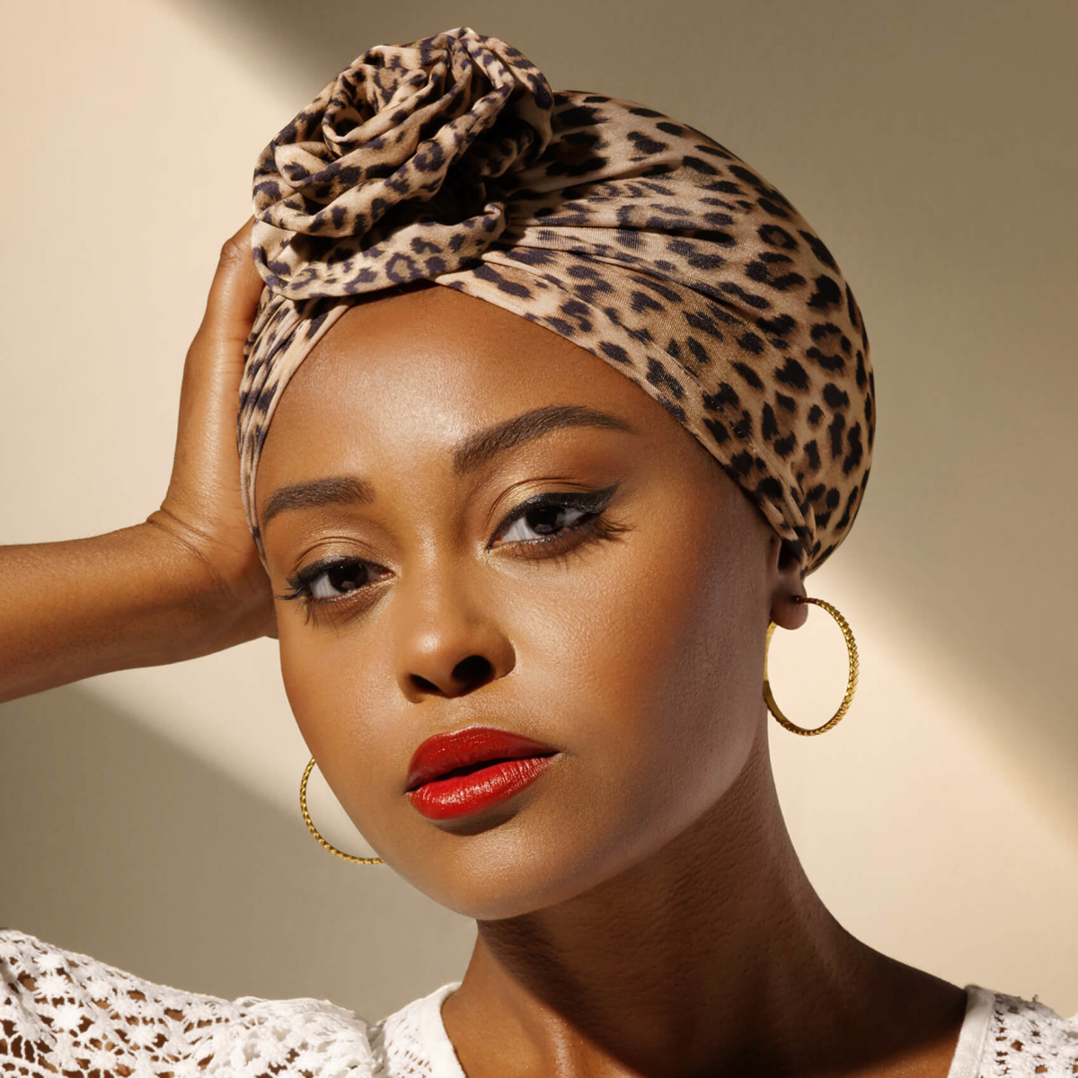 Woman wearing a leopard-print pre-tied turban top knot with a rose detail, gold hoop earrings, red lipstick, and a white lace top against a neutral background.