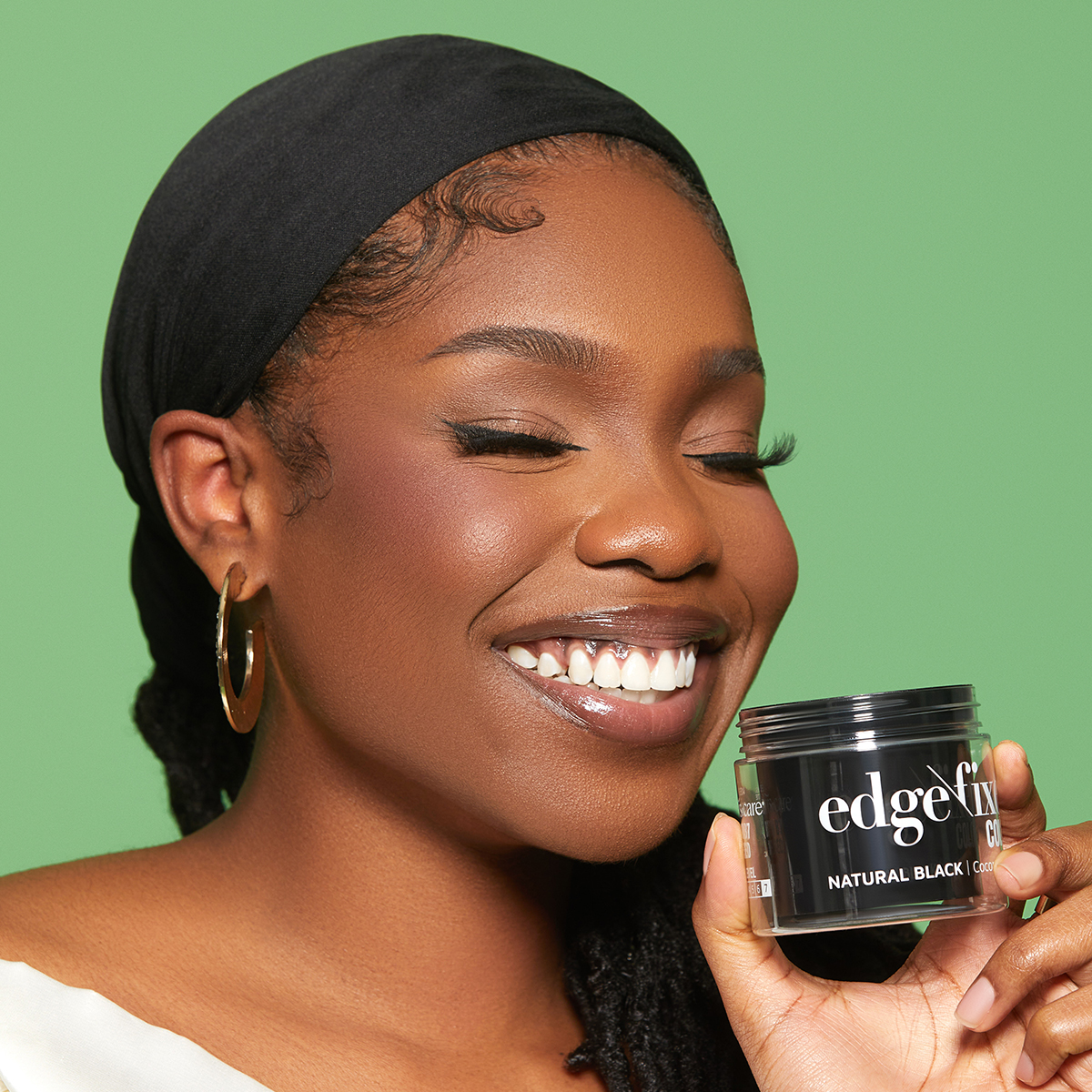 Smiling woman with smooth skin and a black headscarf holds a jar of KISS Colors &amp; Care Edge Fixer in Natural Black against a green background.