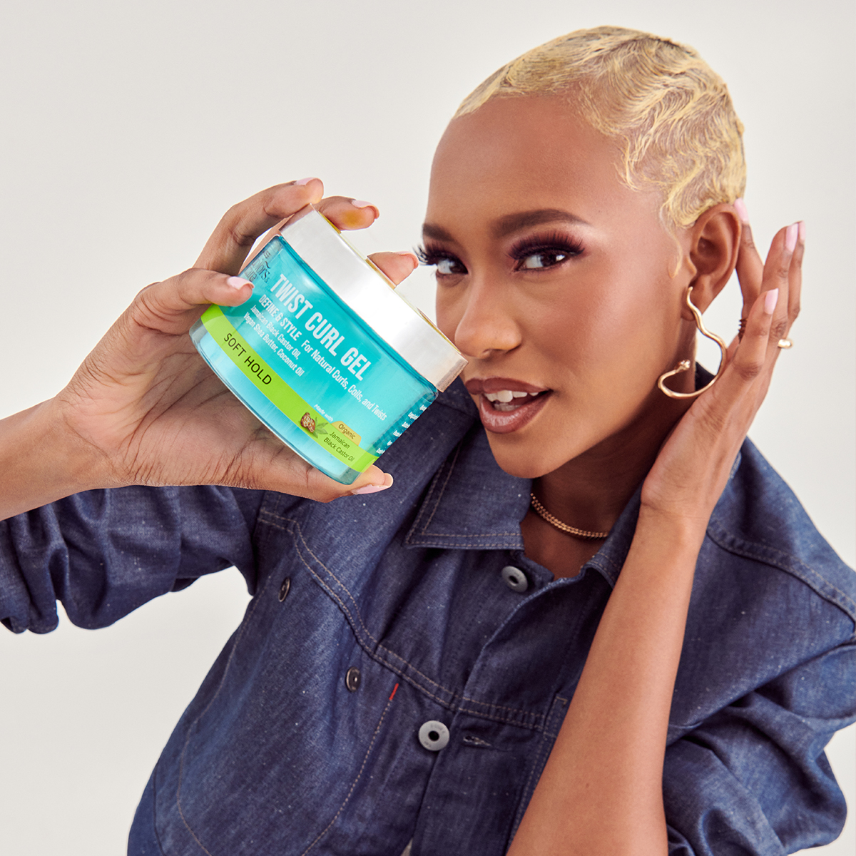 Person with short blonde hair and gold heart-shaped earrings holds a turquoise and green jar of KISS Twist Curl Gel labeled \