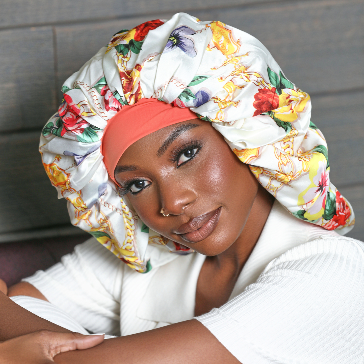 A black woman wearing a super jumbo hair bonnet with a white crepe satin fabric featuring colorful floral and chain patterns, shown in a close-up portrait.