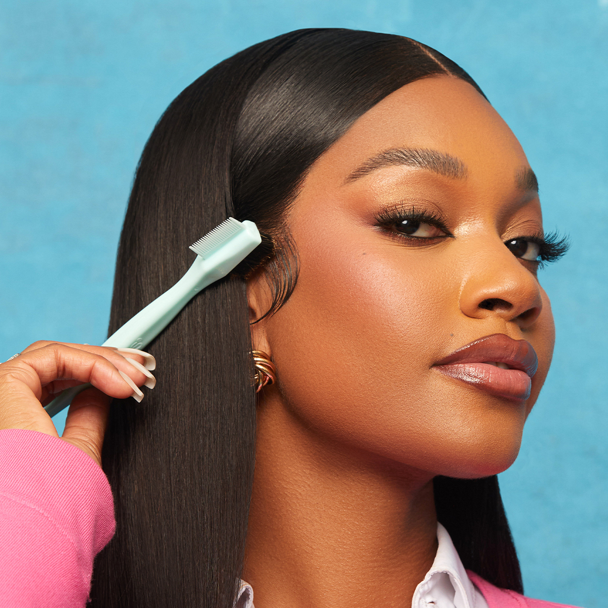 A woman with smooth, straight black hair uses a light mint green boar bristle edge brush to style her baby hairs against a blue background.