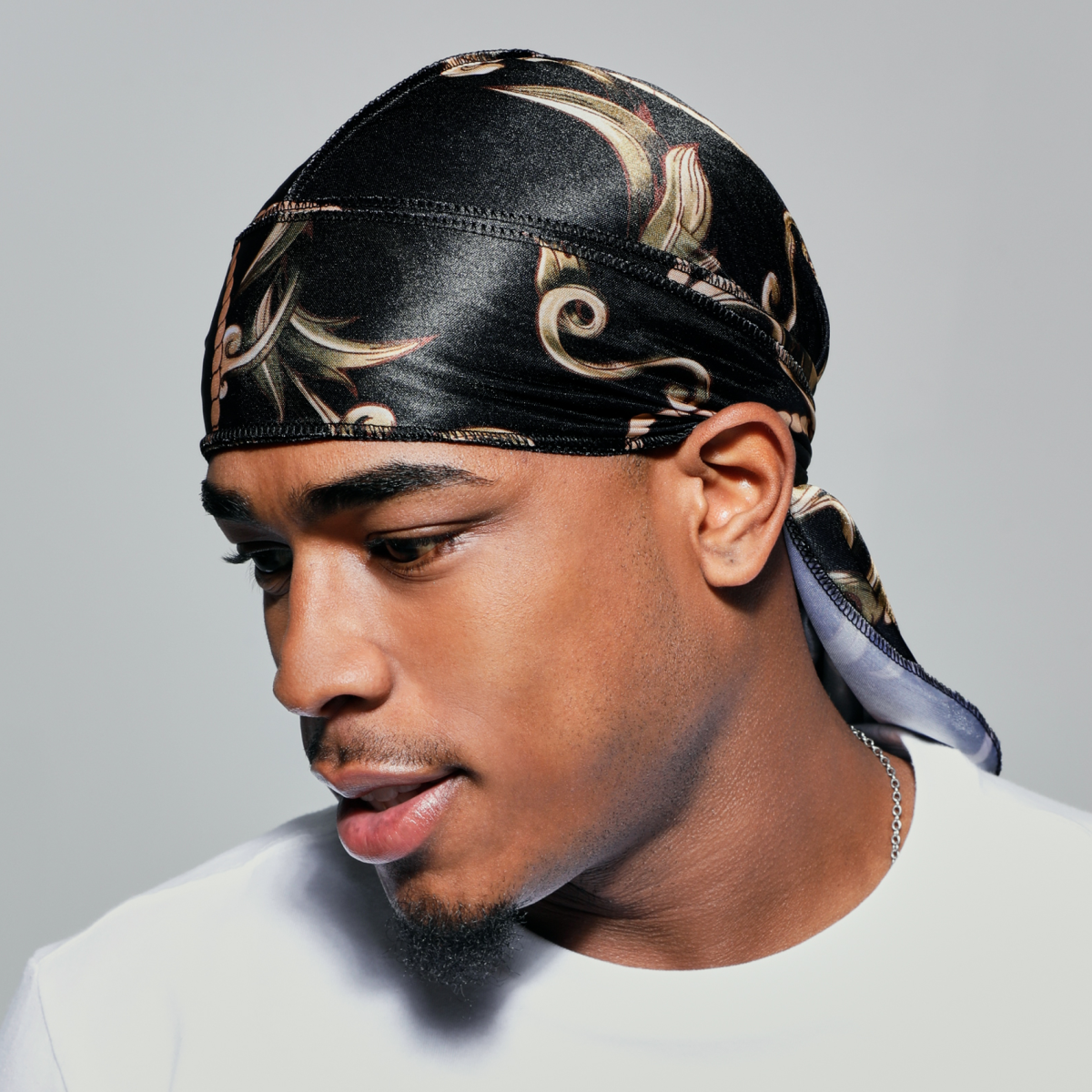 Black silky satin durag with gold and beige intricate swirling pattern, worn by a man facing left, showing side profile and tied at the back.