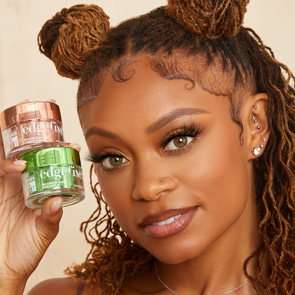 Young woman with styled edges and multiple ear piercings holds two small jars of KISS edge fixer, one rose gold labeled \