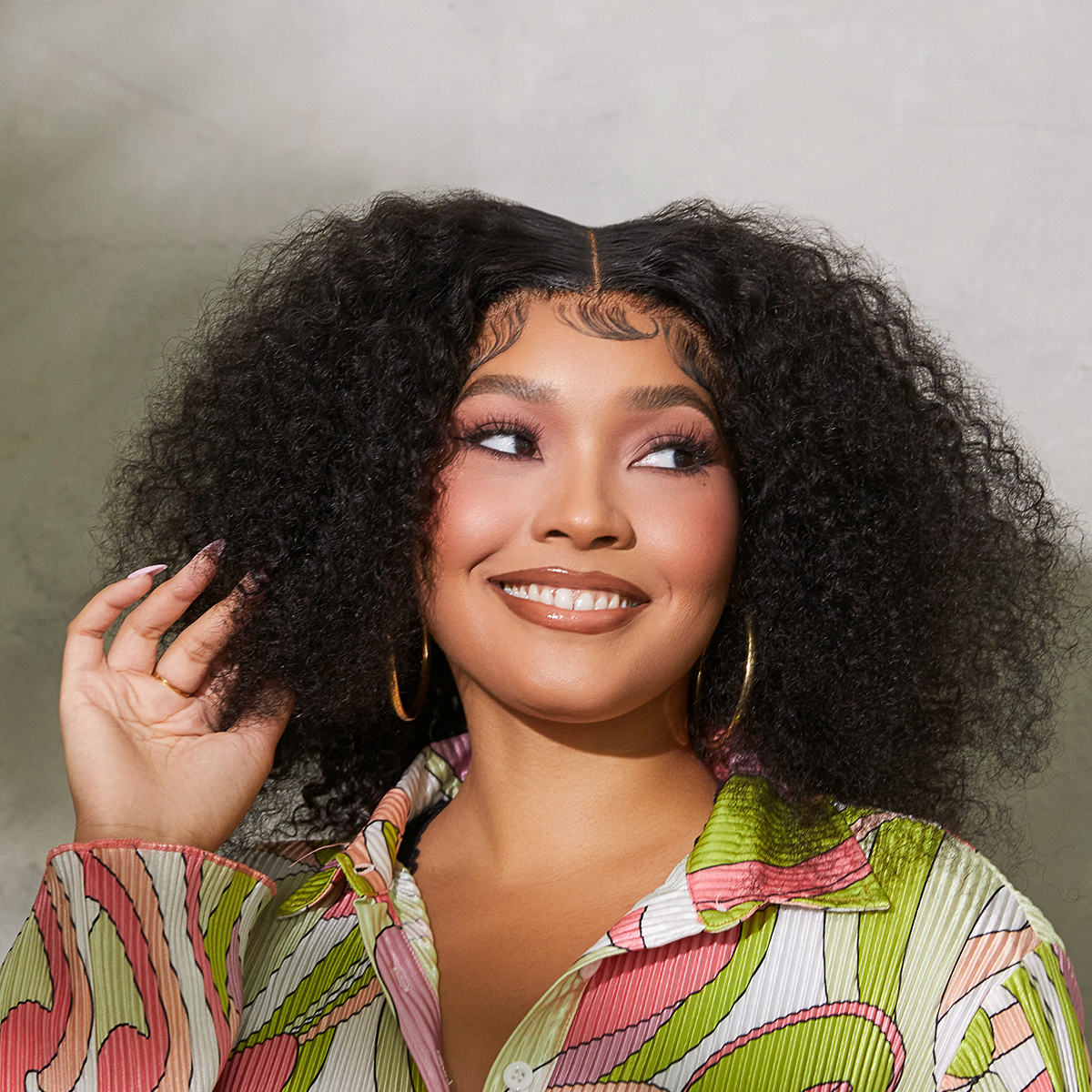 Woman with voluminous medium dark brown curly hair, styled with a center part and defined edges, smiling and wearing gold hoop earrings and a colorful patterned shirt.