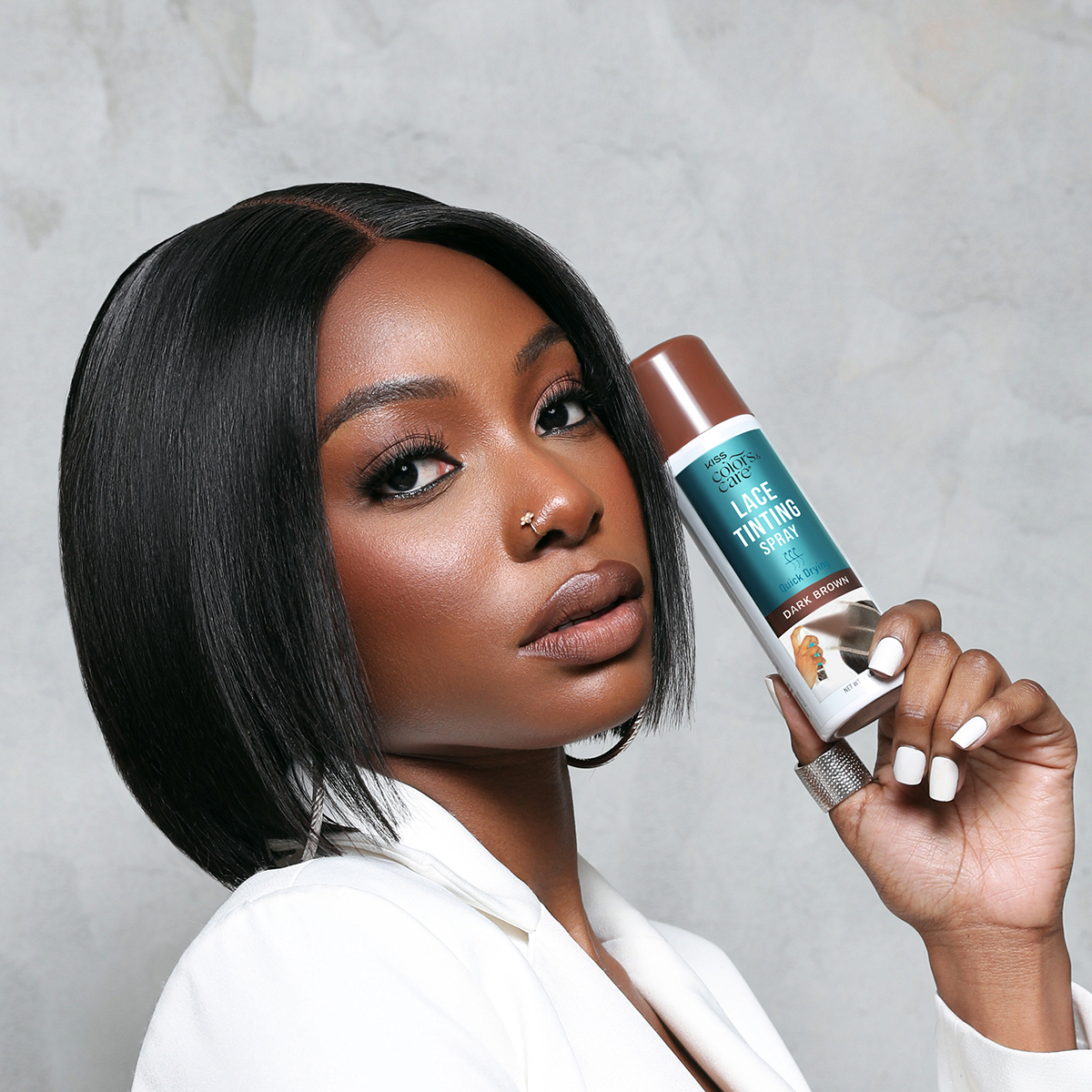 Woman with sleek black bob hairstyle wearing a white blazer holds a can of KISS Colors &amp; Care Lace Tinting Spray in dark brown, labeled quick drying.