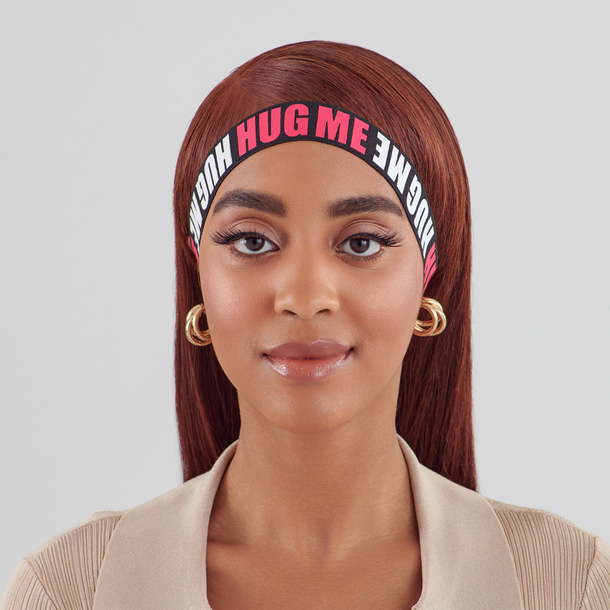 Young woman with straight auburn hair wearing a black elastic band headband with bold red and white text repeating “HUG ME” around her forehead.