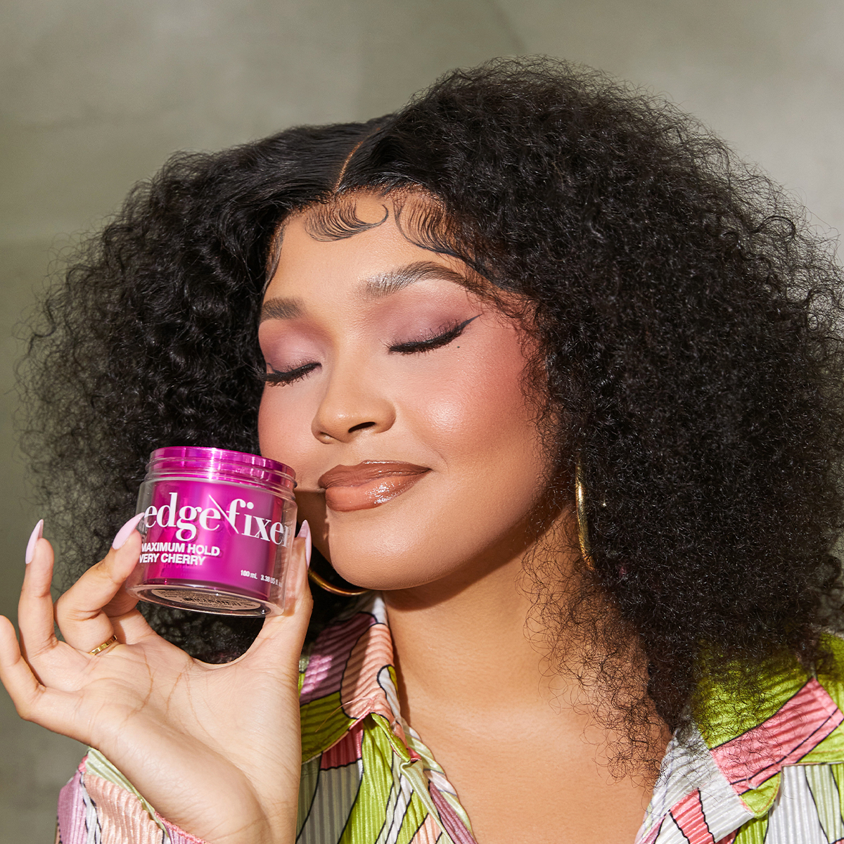 Woman with curly hair and glossy makeup holding a pink jar of KISS Colors &amp; Care Edge Fixer labeled \