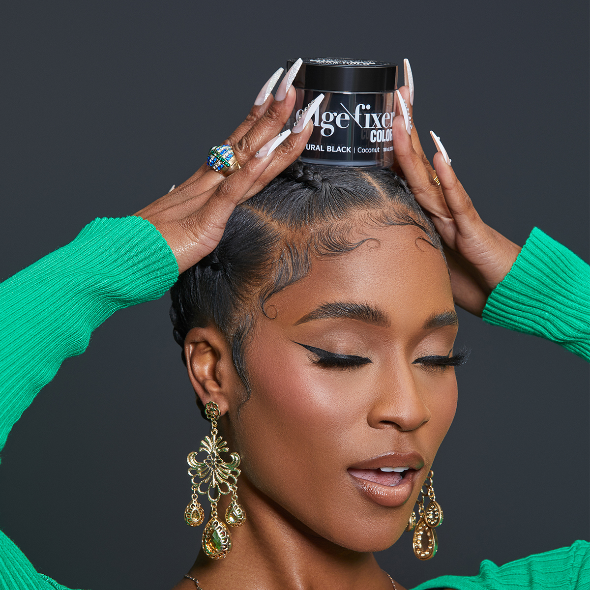 Woman with braided hair and winged eyeliner wearing gold chandelier earrings and green ribbed sleeves holds a jar of KISS Age Fixer Edge Color in Natural Black on her head.