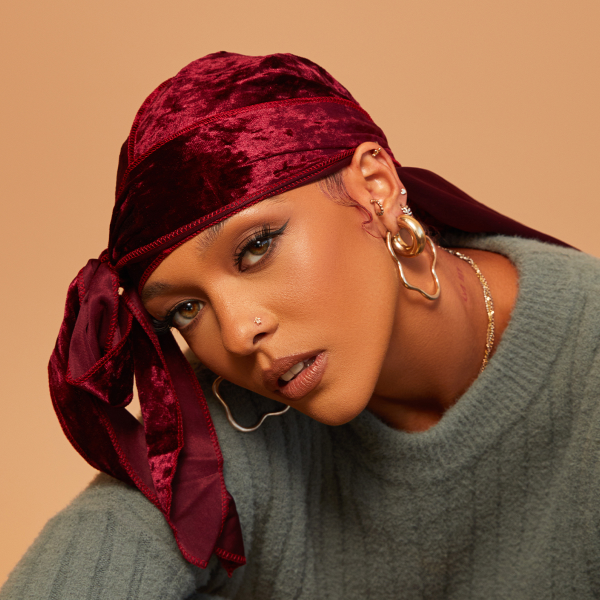 Close-up of a person wearing a burgundy velvet durag tied in a bow on the side, paired with gold hoop earrings, a small nose stud, and a textured gray-green sweater against a beige background.
