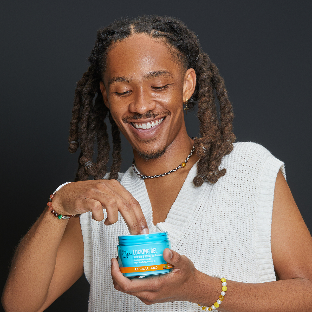 Smiling person with long dreadlocks wearing a white sleeveless sweater dips finger into a blue jar of KISS Colors &amp; Care Locking Gel labeled for maintaining and defining with regular hold.
