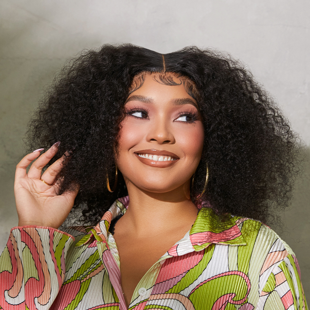 Young woman with voluminous curly black hair, wearing a colorful patterned shirt and gold hoop earrings, smiling with makeup in a neutral-toned background.