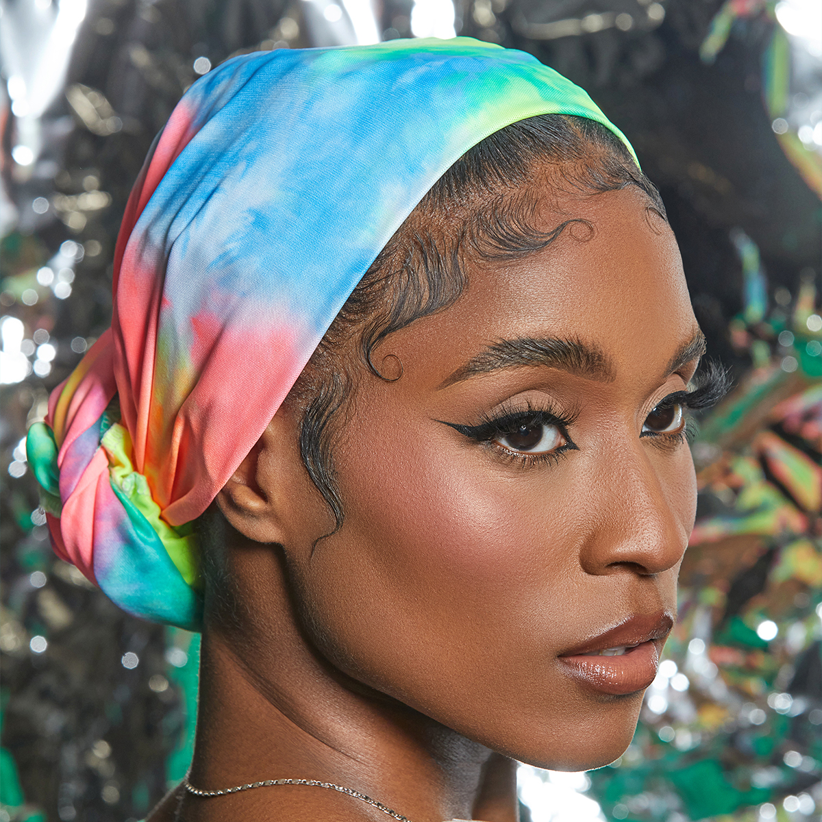 Woman shown in close-up wearing a soft satin wrap scarf with vibrant tie-dye colors of blue, green, yellow, and pink, styled as a head wrap against a shiny, reflective background.