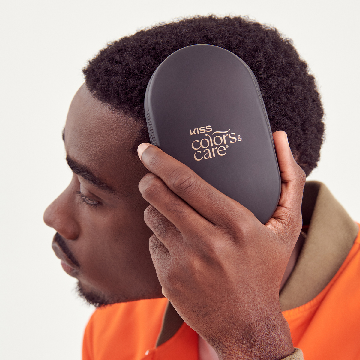 Man with short curly black hair wearing an orange jacket holds a matte black KISS Colors &amp; Care hairbrush to the side of his head.