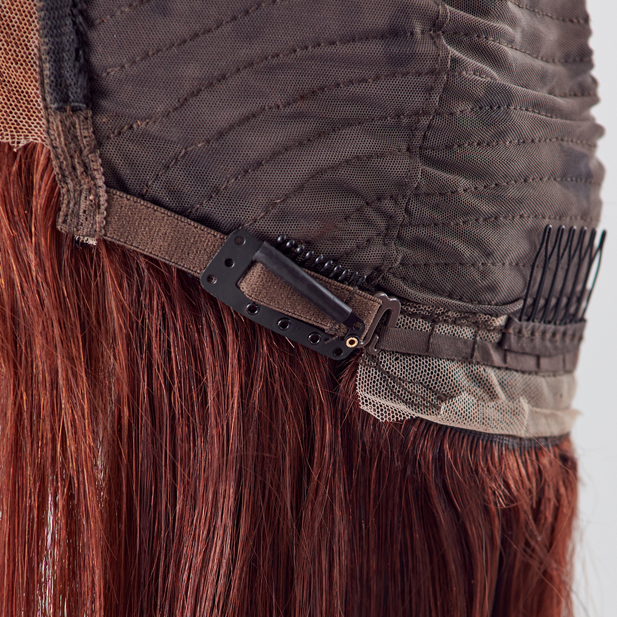 Close-up of the inside of a brown lace wig cap showing attached black snap-on wig clips and a comb for secure fit, with reddish-brown hair strands.