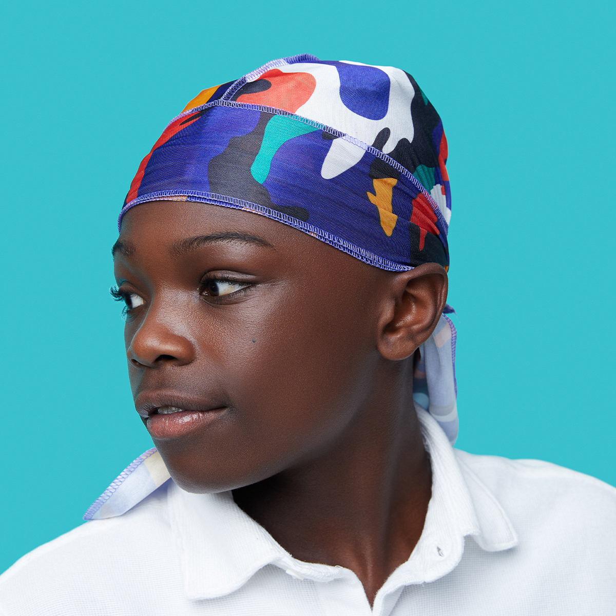 Young person wearing a multicolored camouflage satin durag with blue, purple, red, white, and yellow patterns, against a solid turquoise background.