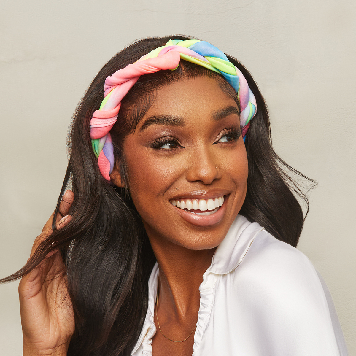 Woman smiling and looking to the side, wearing a twisted soft satin scarf wrap in pastel rainbow colors over dark wavy hair, dressed in a white ruffled blouse.