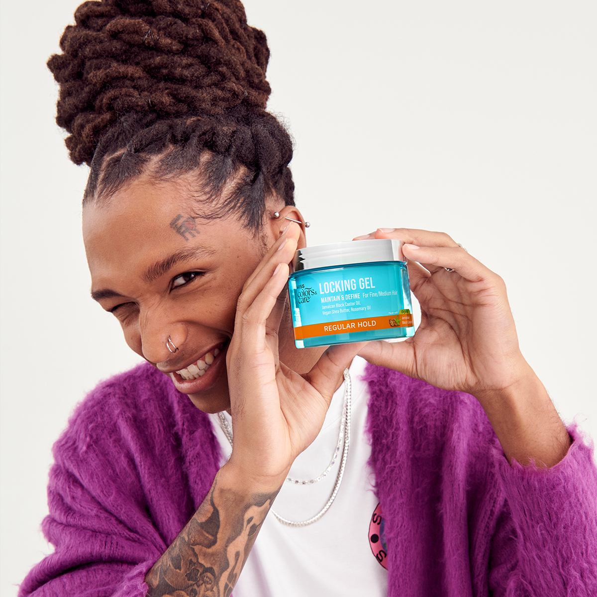 Person with braided hair and tattoos smiles while holding a blue jar of KISS Colors &amp; Care Locking Gel, labeled for fine/medium hair with regular hold.