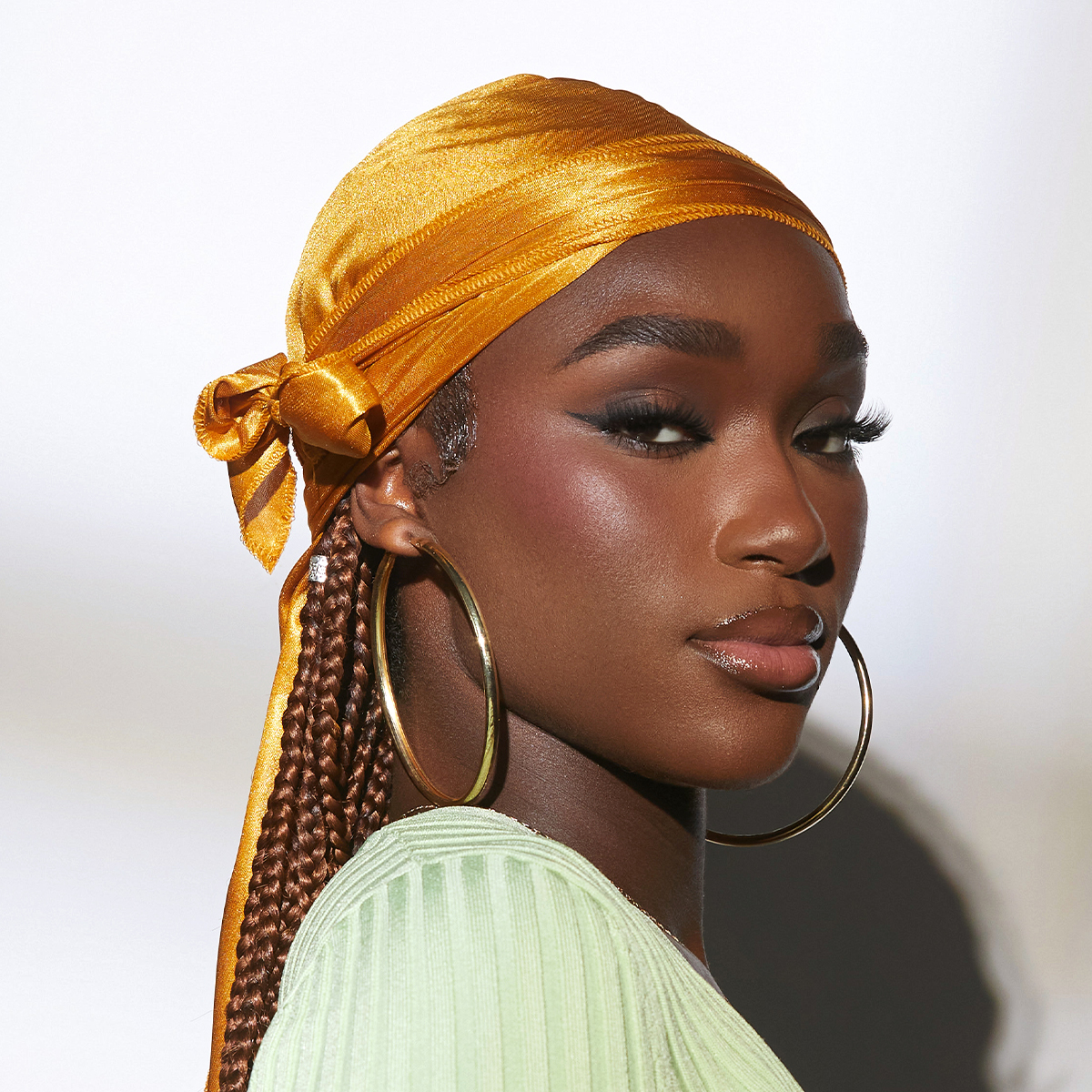 Shiny gold satin durag tied at the back worn by a woman with braided hair, large gold hoop earrings, and a light green ribbed top against a plain background.