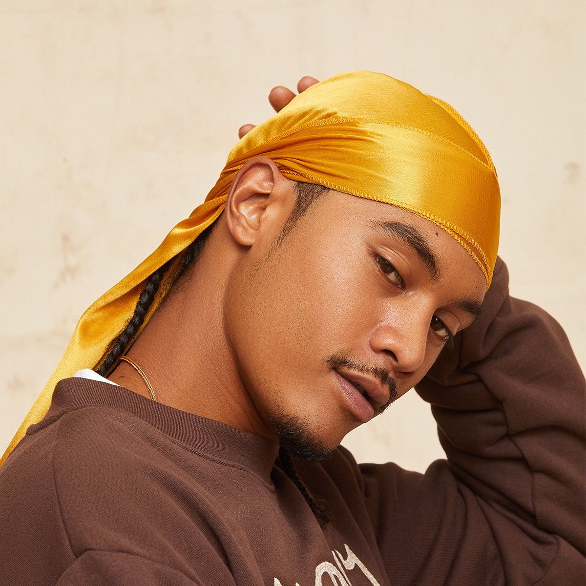 Man wearing a shiny gold satin durag tied at the back, shown in close-up side profile against a neutral background, wearing a brown sweatshirt.