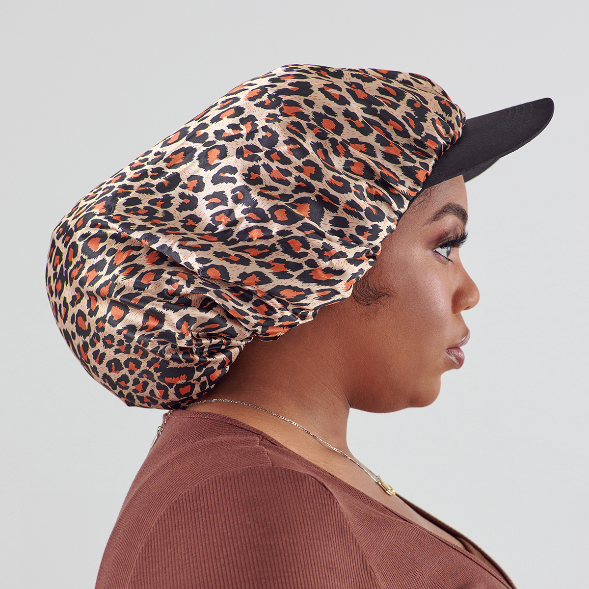 Woman shown in profile wearing an XL silky satin bonnet cap with a leopard print and a black visor, paired with a brown top and silver necklace.