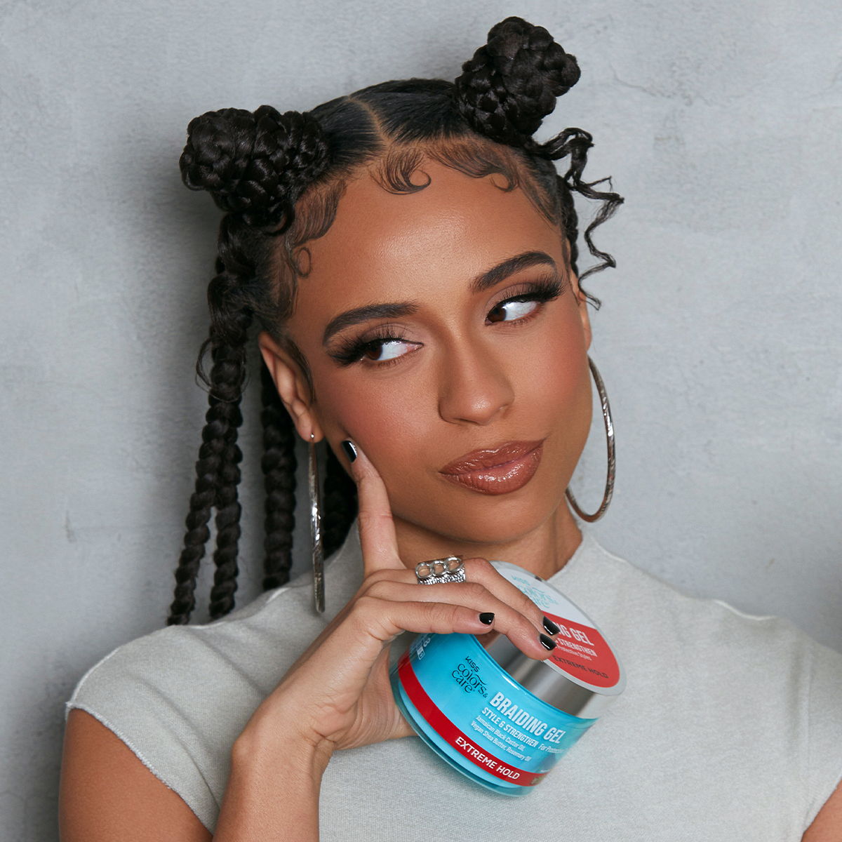 Woman with braided hair styled in two buns and baby hairs, wearing large hoop earrings and a light gray sleeveless top, holding a blue and silver container of KISS Colors &amp; Care Braiding Gel with a red label stating \
