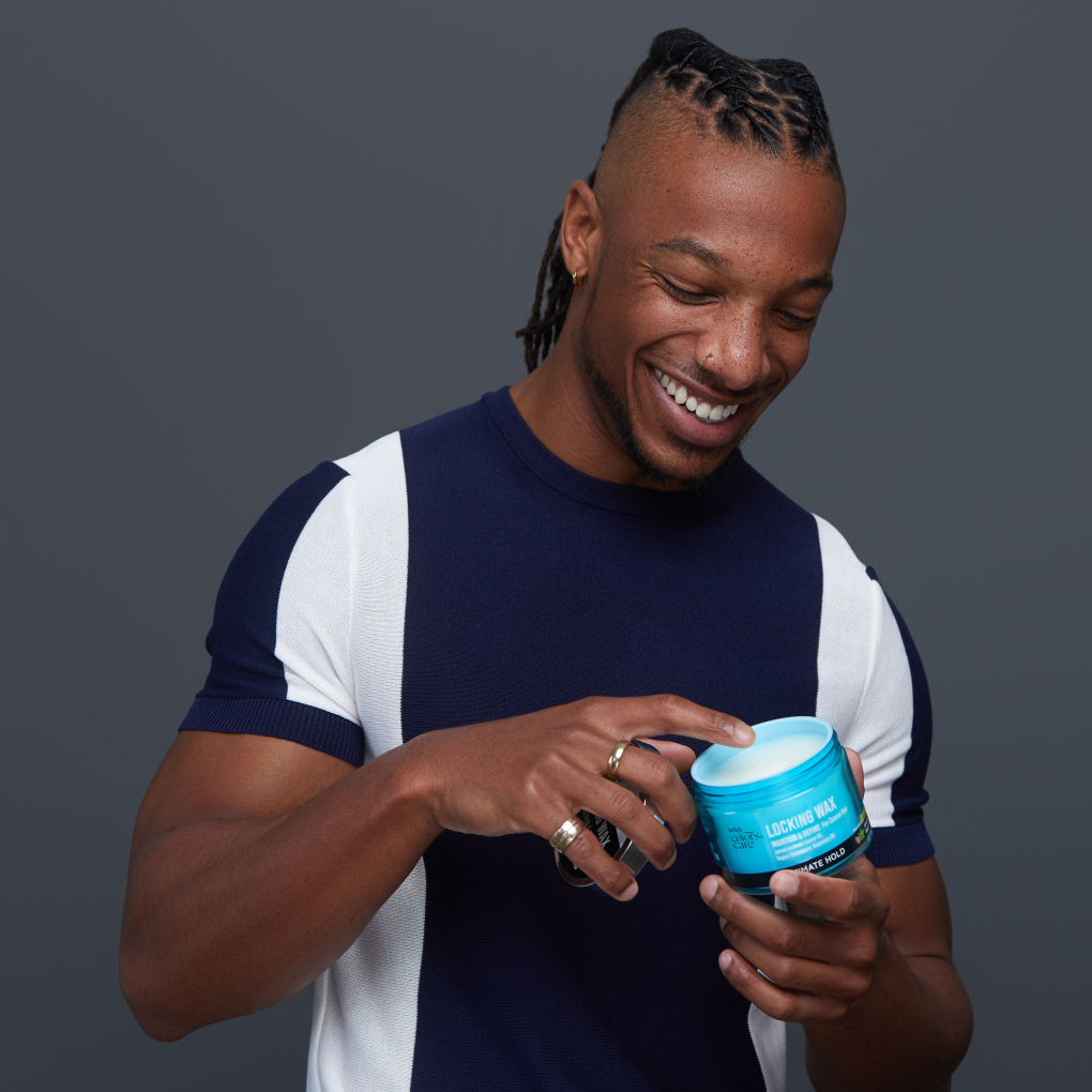 Man wearing a navy and white short-sleeve shirt smiling while holding a blue jar of KISS Colors &amp; Care Locking Wax with the lid off, finger touching the wax inside.