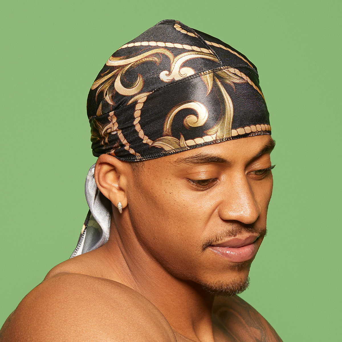 Man wearing a silky satin durag with black and gold intricate wave and scroll patterns, shown in profile against a green background.