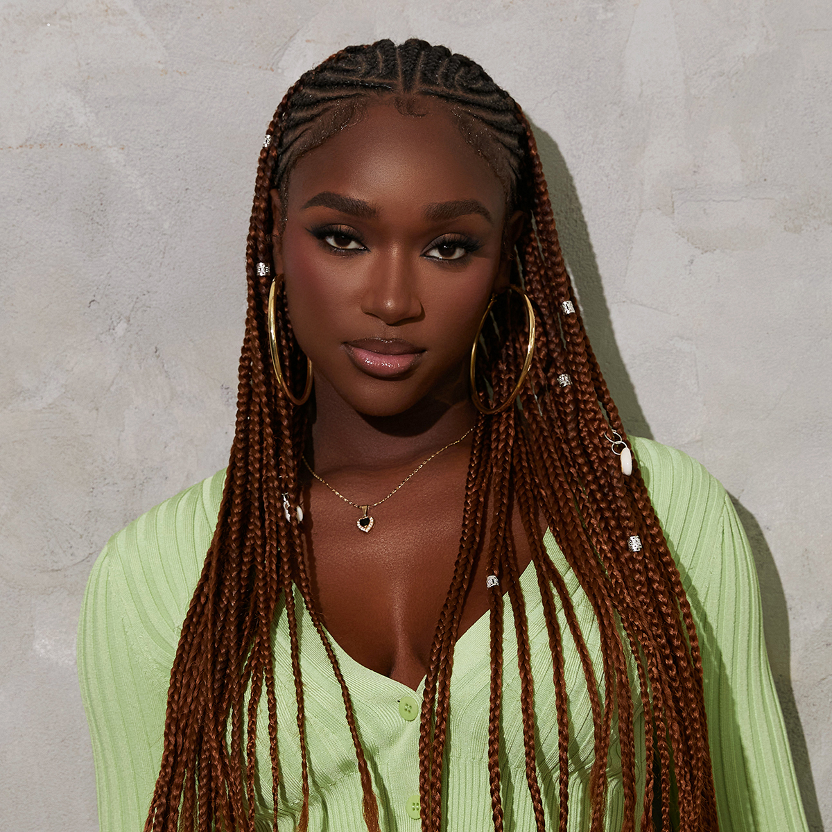 Dark-skinned woman with long brown box braids adorned with small silver braid charms, wearing large gold hoop earrings and a light green ribbed cardigan.