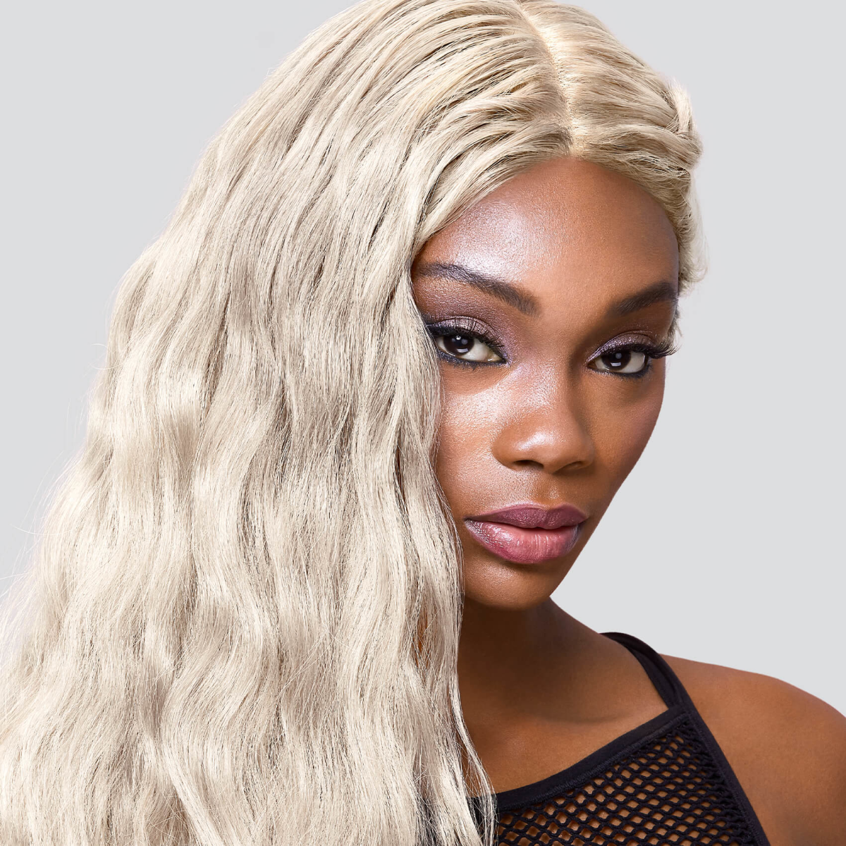 Close-up of a woman with long, wavy platinum blonde hair styled to the side, wearing a black mesh top against a light gray background.
