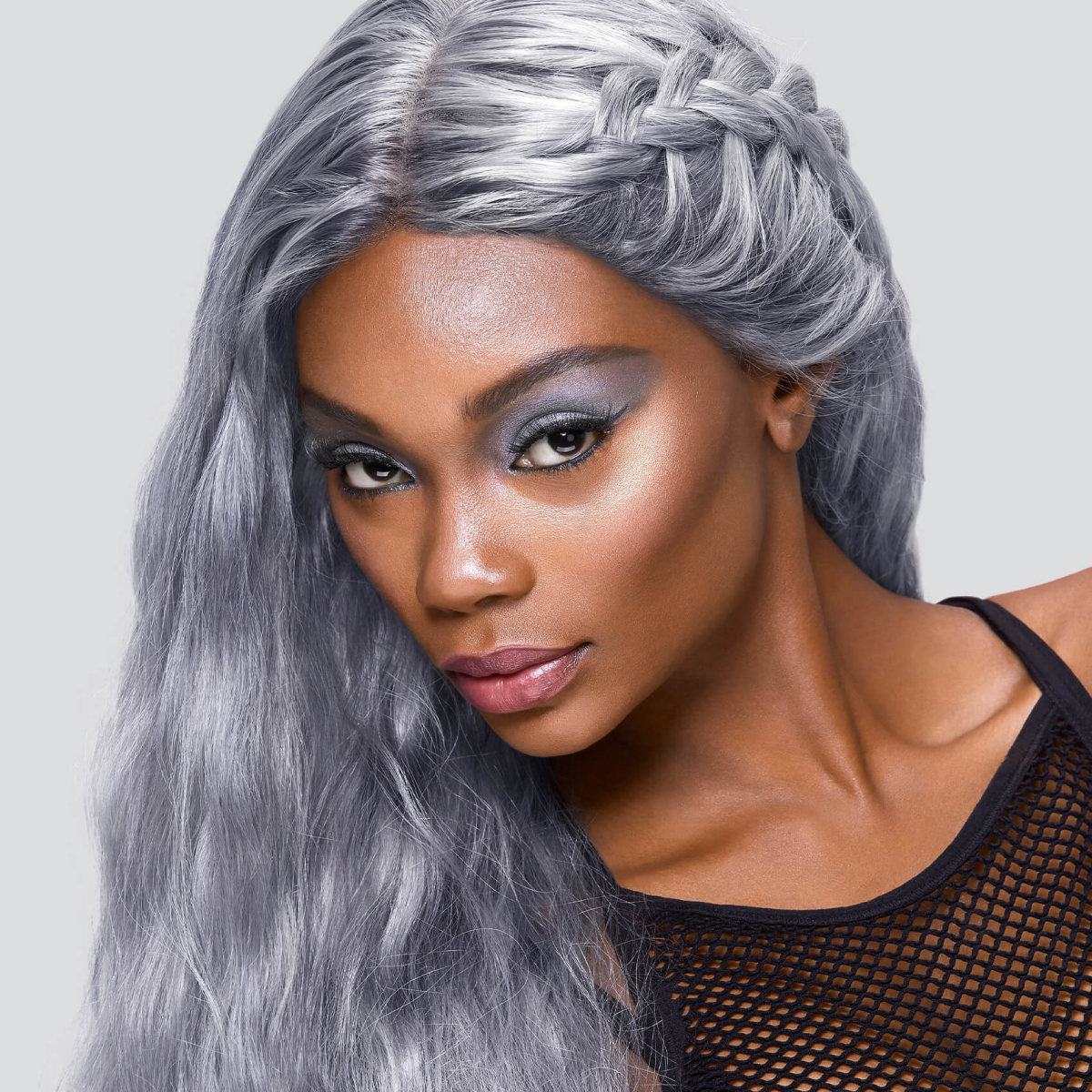 Close-up of a woman with long, braided silver-gray hair styled in loose waves, wearing a black mesh top and cool-toned smoky eye makeup, showcasing the ice-toned bleach and tone hair color.