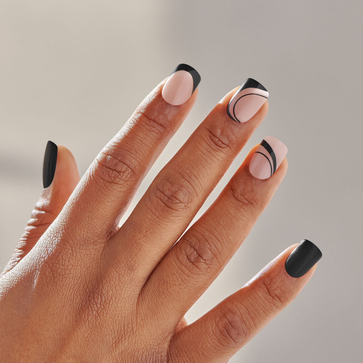 Hand with medium brown skin tone wearing press-on manicure nails in a wide fit. Nails feature a black dress design with matte black tips, blush pink base, and curved black lines on some nails.