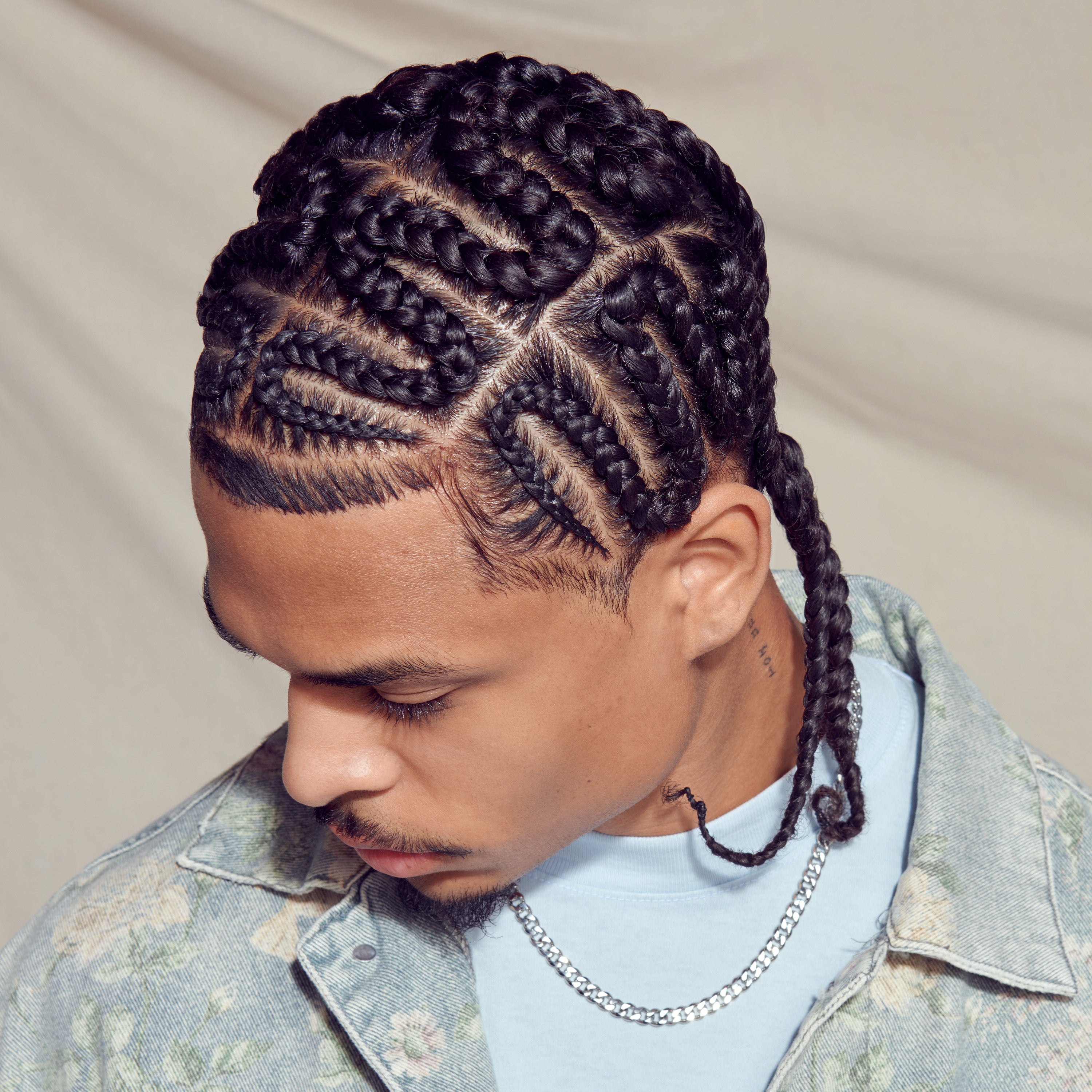 Man with dark braided hair styled in cornrows looking down, wearing a light blue shirt and a floral-patterned denim jacket with a silver chain necklace.