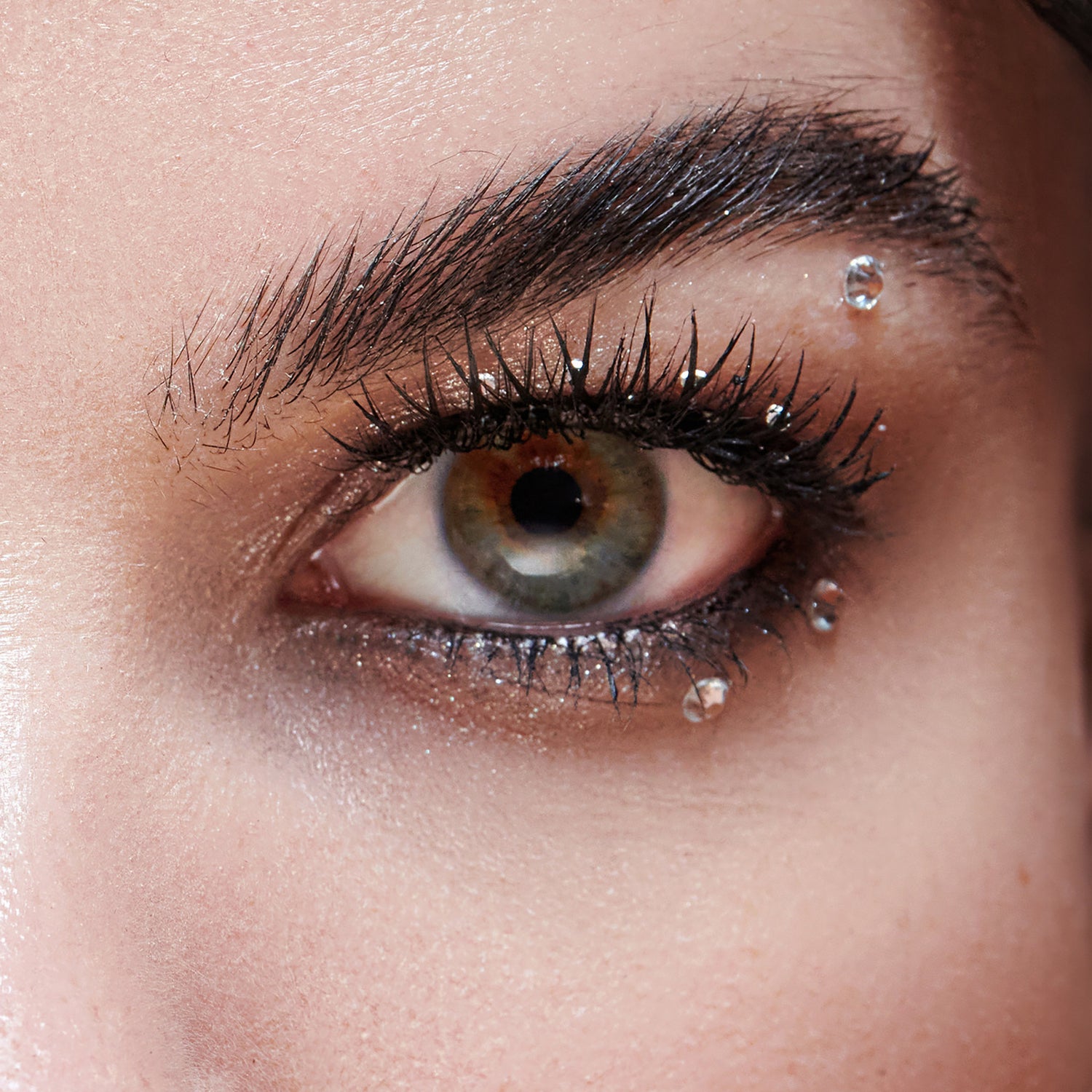 The image features a close-up of a human eye, showcasing striking details. The eye has a vibrant, multi-colored iris with shades of green and brown. Long, dark eyelashes frame the eye, enhancing its allure. A small, sparkling droplet of water or a decorative gem is positioned just below the eye, adding a touch of glamour. The skin around the eye appears smooth, with a subtle shimmer, contributing to an overall captivating and artistic look.