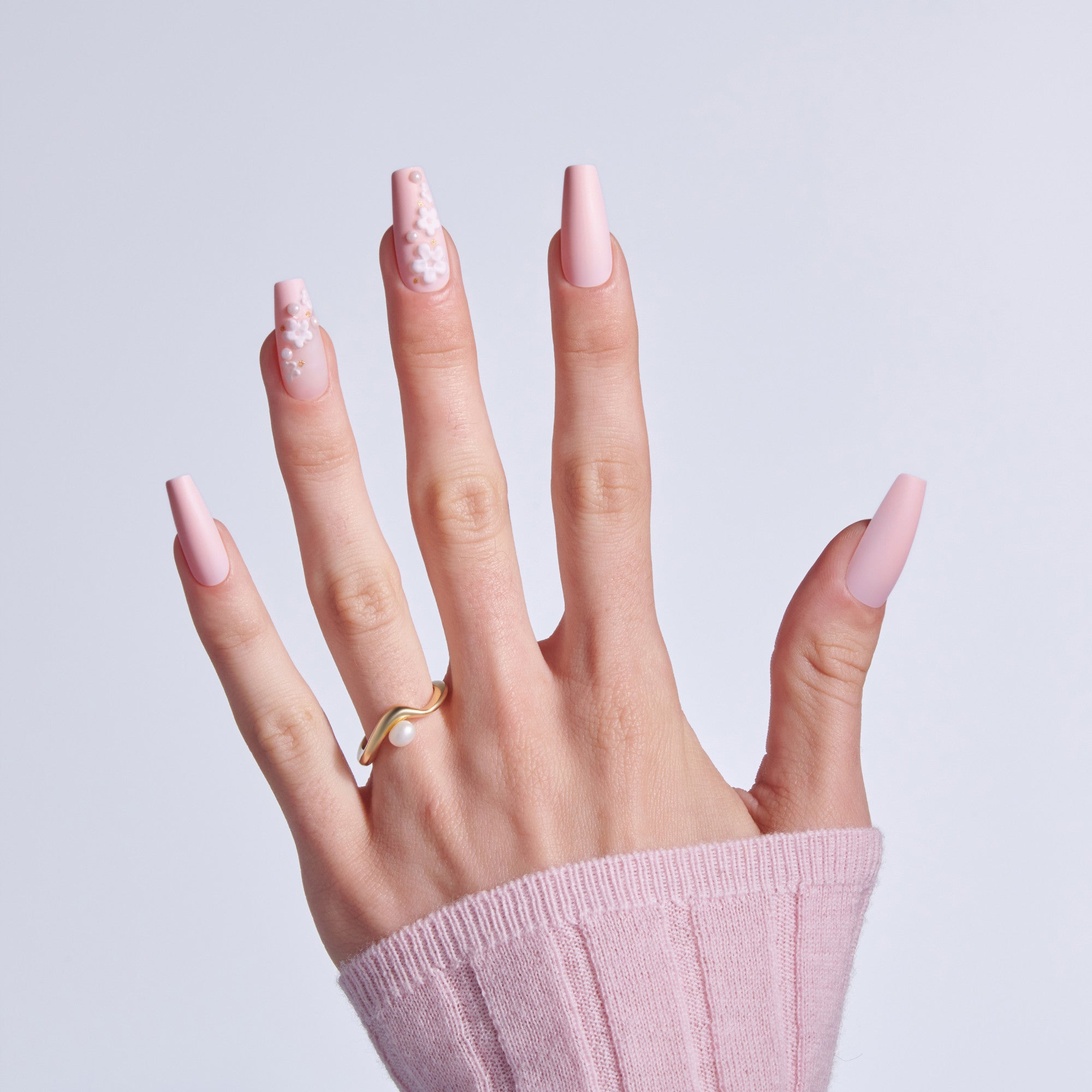 Hand with pink coffin nails, some decorated with white floral design, wearing gold pearl ring.