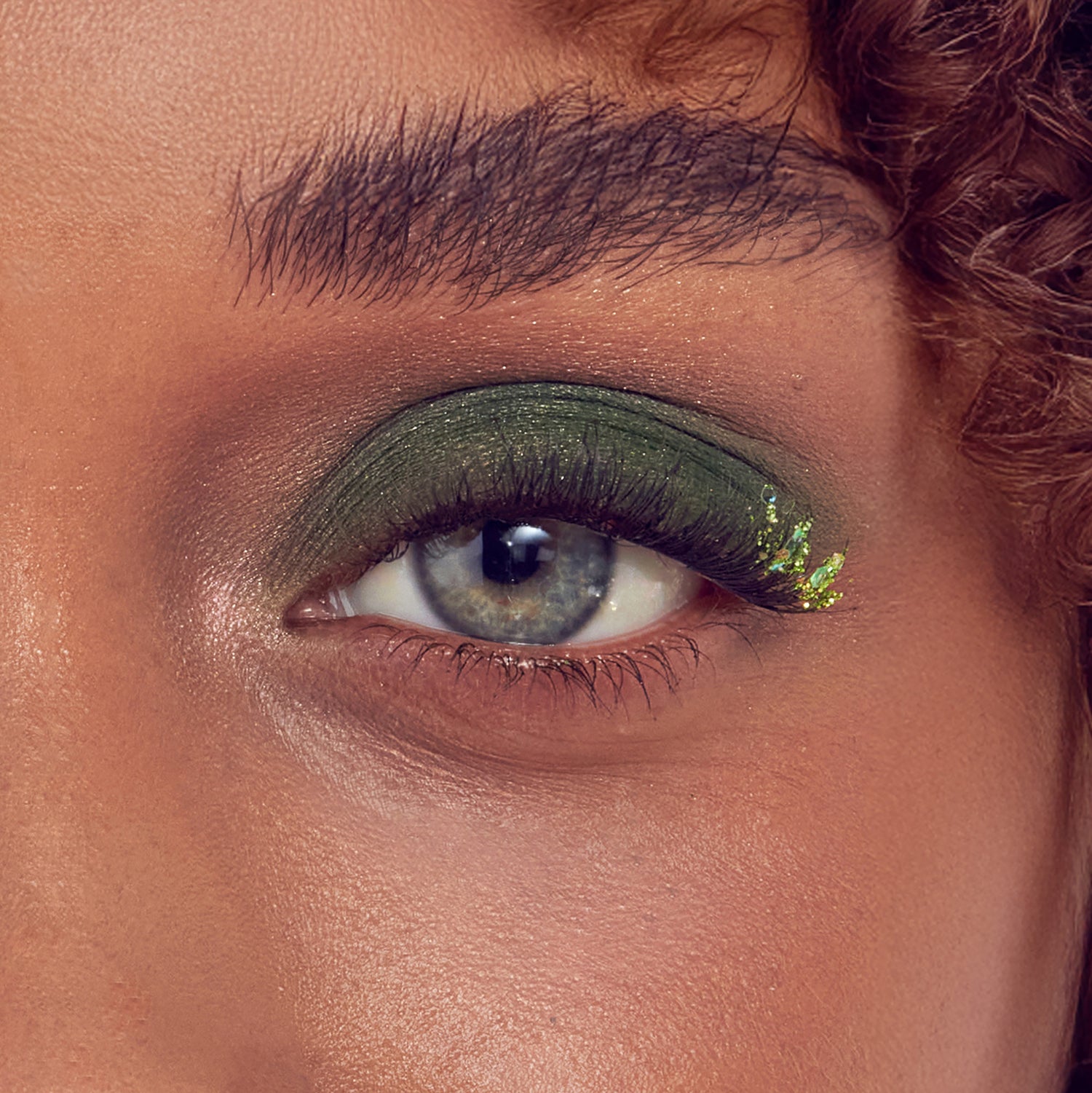 Close-up of eye with green eyeshadow, gold glitter, and black eyeliner on warm-toned skin.