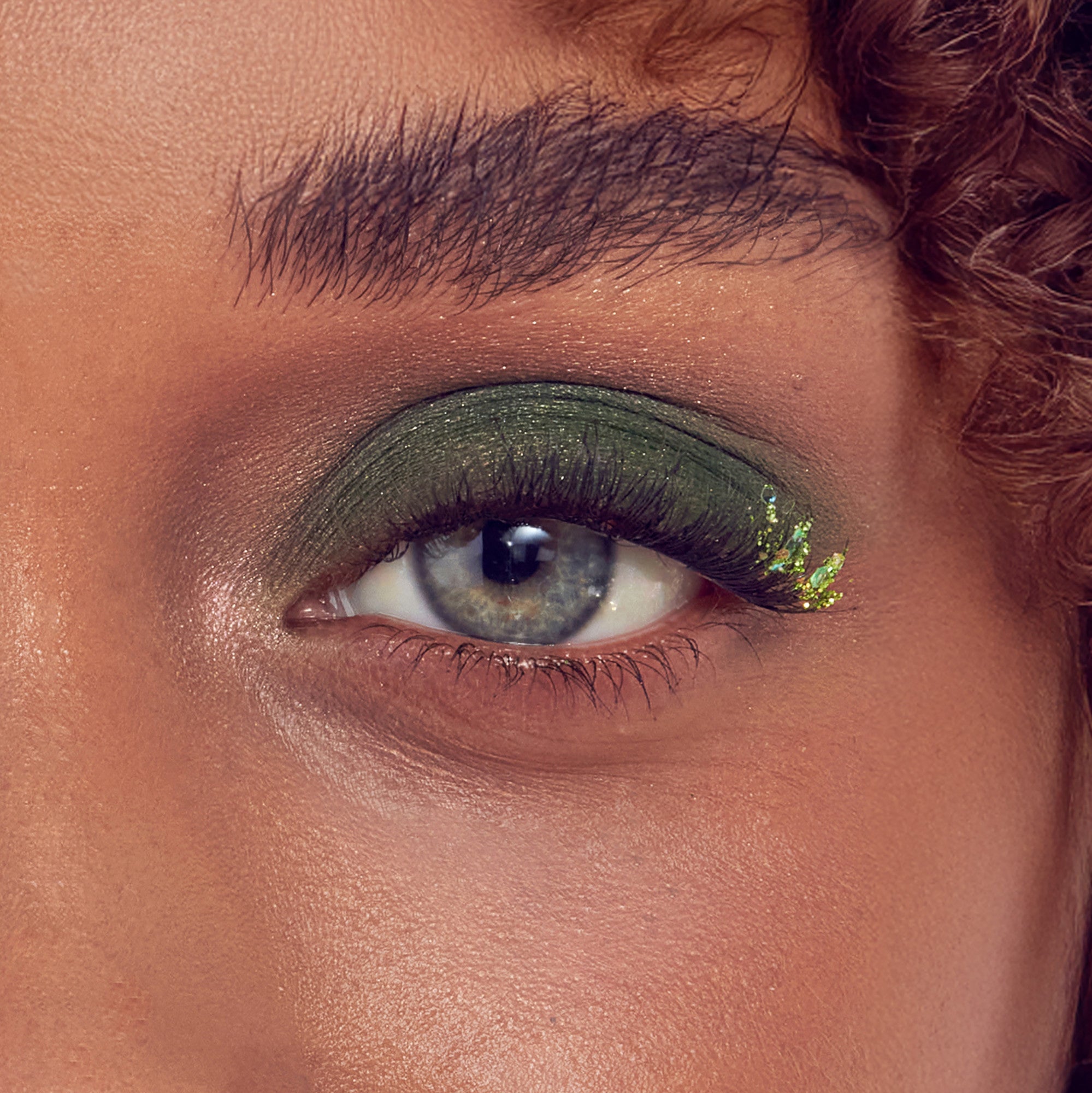 Close-up of eye with green eyeshadow, gold glitter, and black eyeliner on warm-toned skin.