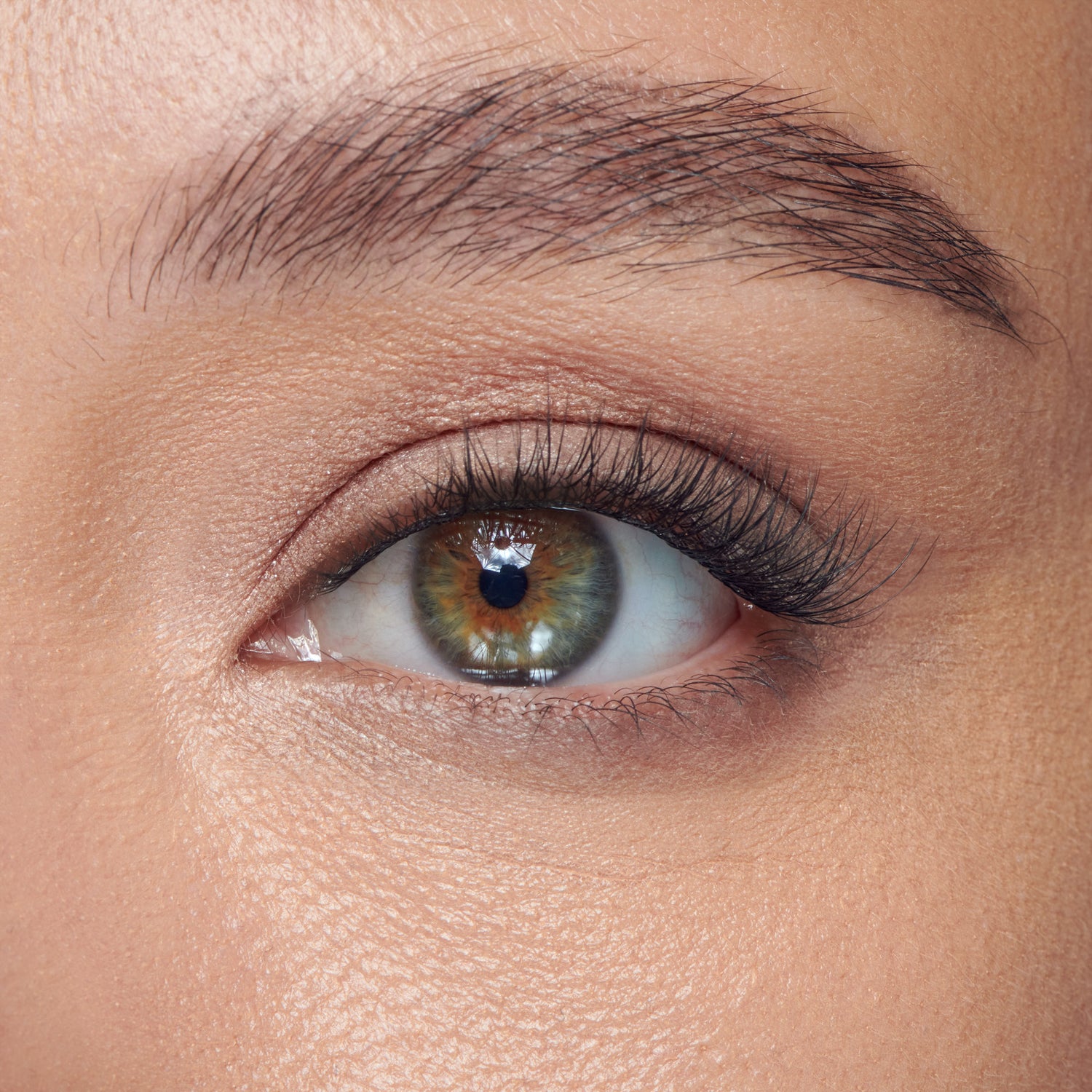 The image features a close-up of a human eye, showcasing a striking combination of colors in the iris, which includes shades of green and hints of brown. The eye is framed by well-groomed eyebrows and has long, dark eyelashes. The skin around the eye appears smooth and lightly textured, highlighting the natural beauty of the eye. The overall focus is on the intricate details of the eye, emphasizing its depth and clarity.