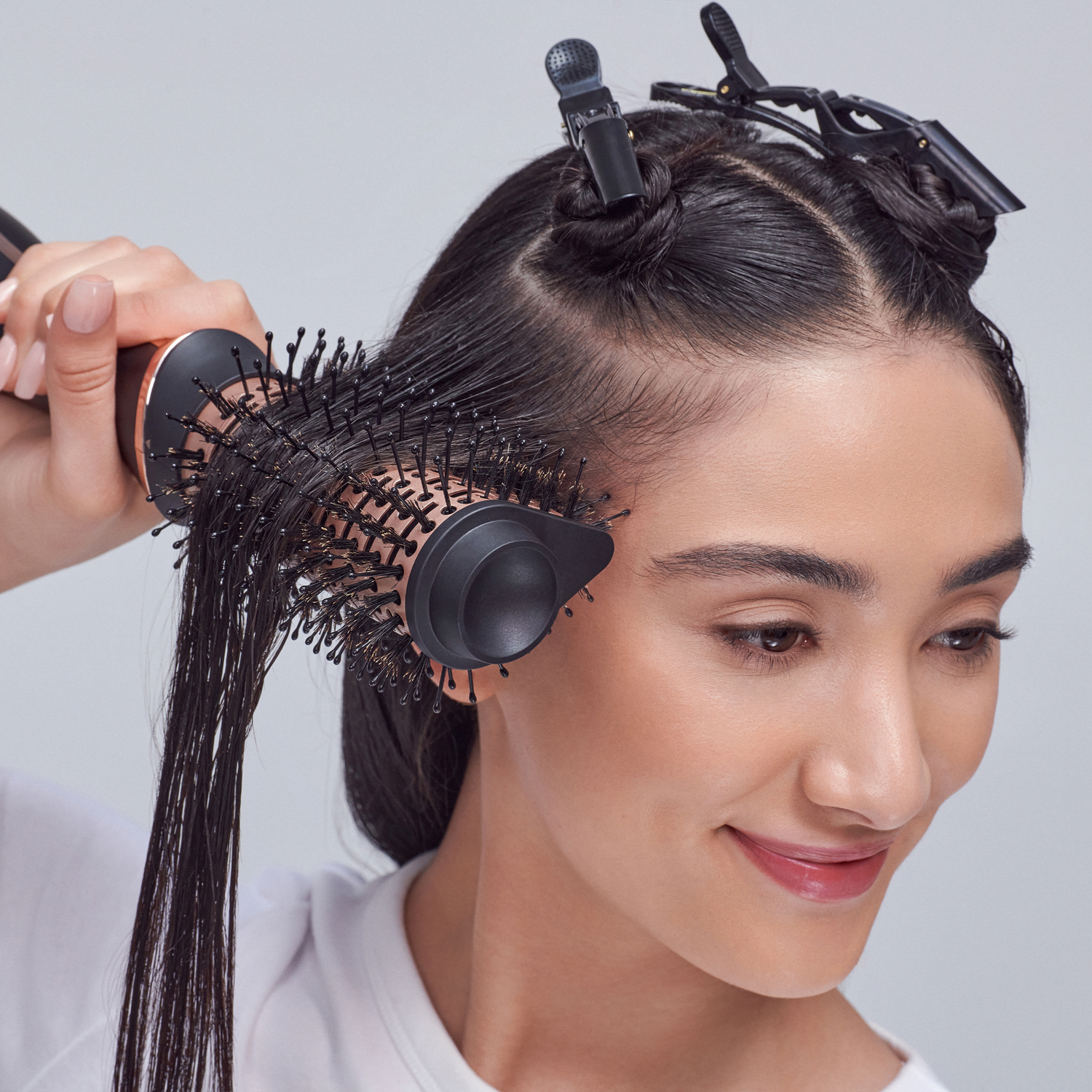 The image features a woman with long, straight hair being styled. She is using a round brush with bristles to smooth and shape her hair. Her hair is partially sectioned and clipped back, indicating that she is in the process of styling. The background is neutral, which helps to focus on her and the styling tools. The woman has a pleasant expression, suggesting she is enjoying the hair styling process.