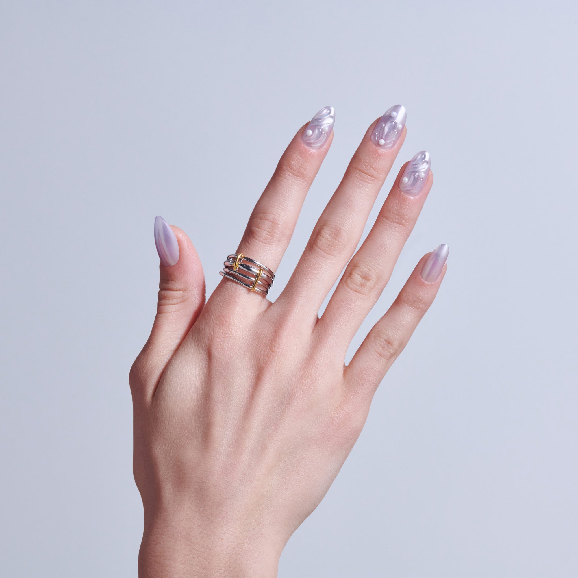 Hand with translucent lavender nail art and gold stacked rings on light blue background