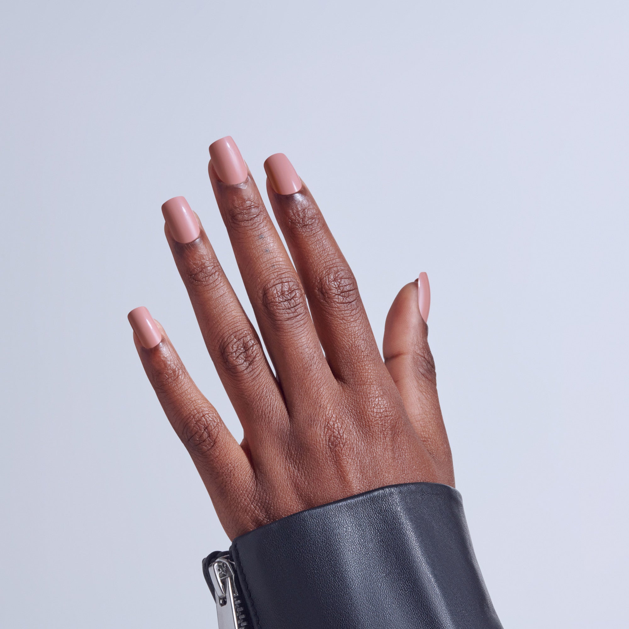 Hand with nude pink ombre nail manicure and dark leather cuff against light background.