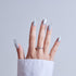 Hand with silver glitter acrylic nails and ornate silver ring against gray background.