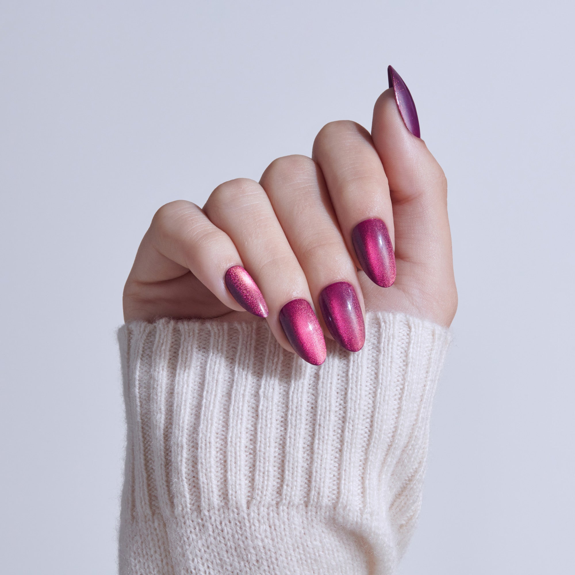 Manicured hand with almond-shaped nails in magenta shimmer polish, wearing cream sweater.