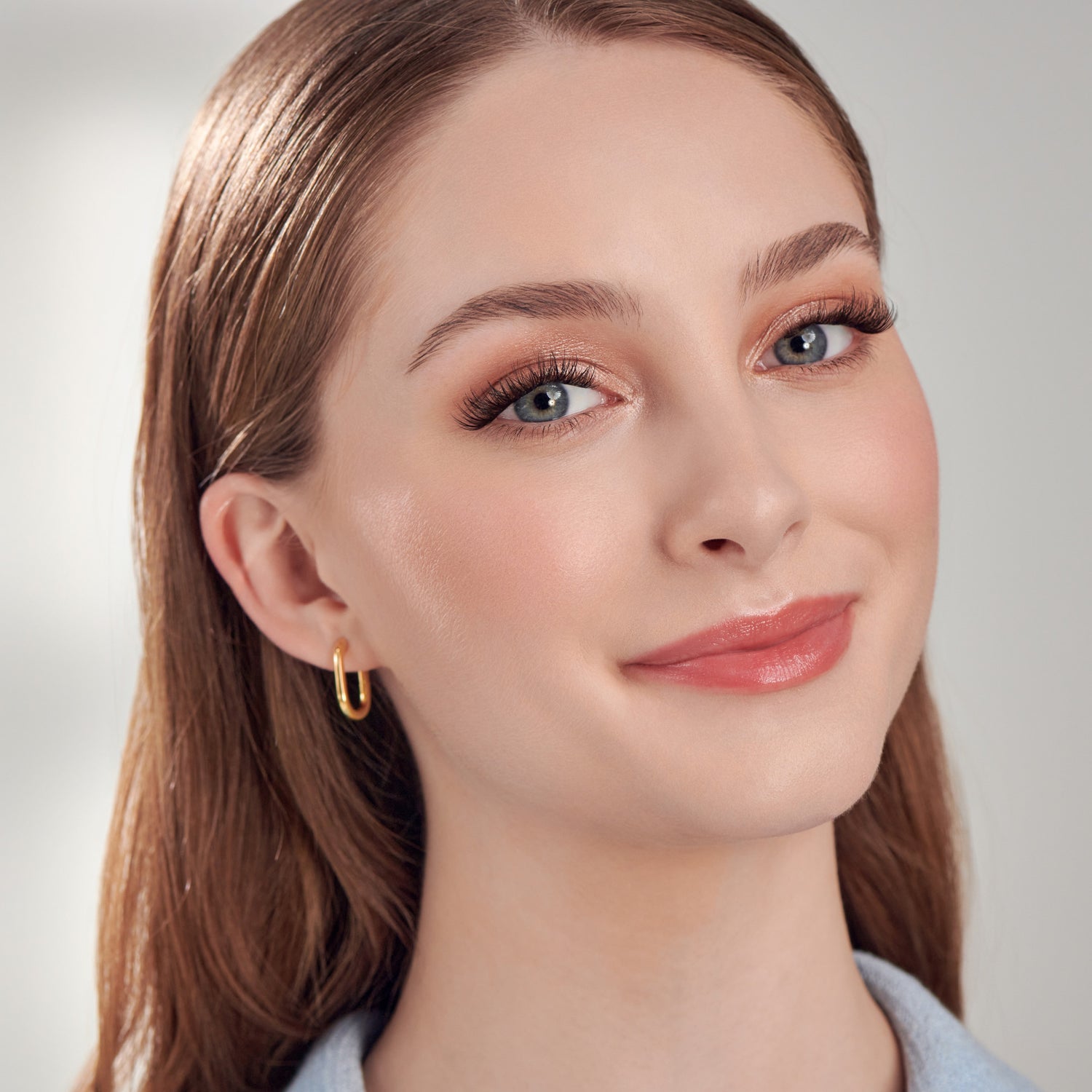 The image features a young woman with long, straight hair and a warm smile. She has a natural makeup look, highlighting her eyes with subtle eyeshadow and long eyelashes. Her lips are painted in a soft, peachy shade. She is wearing a pair of simple gold hoop earrings, which add a touch of elegance to her appearance. The background is softly blurred, creating a clean and professional look.