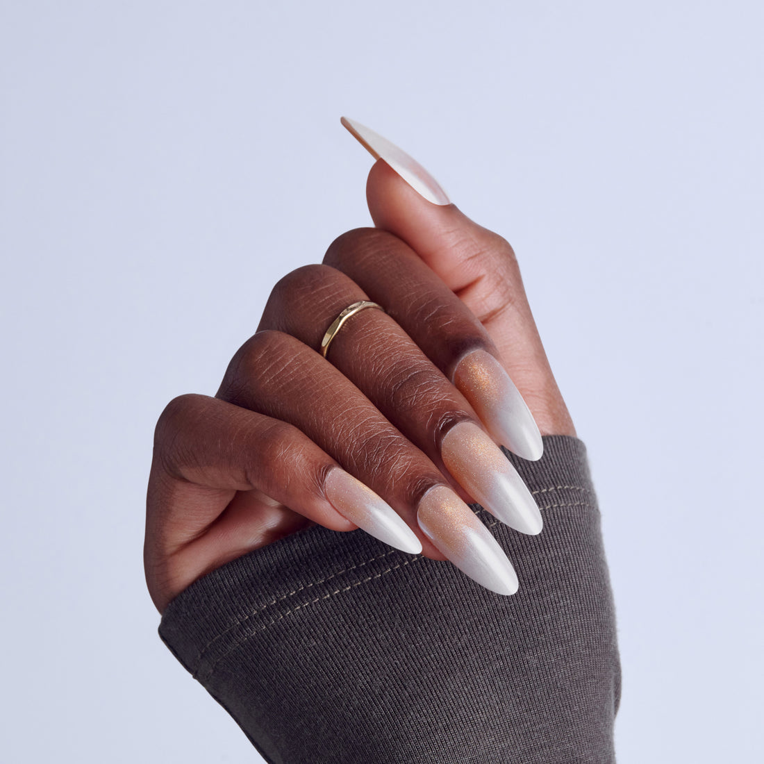 Close-up of manicured hand with white ombre almond nails and gold ring against light background.