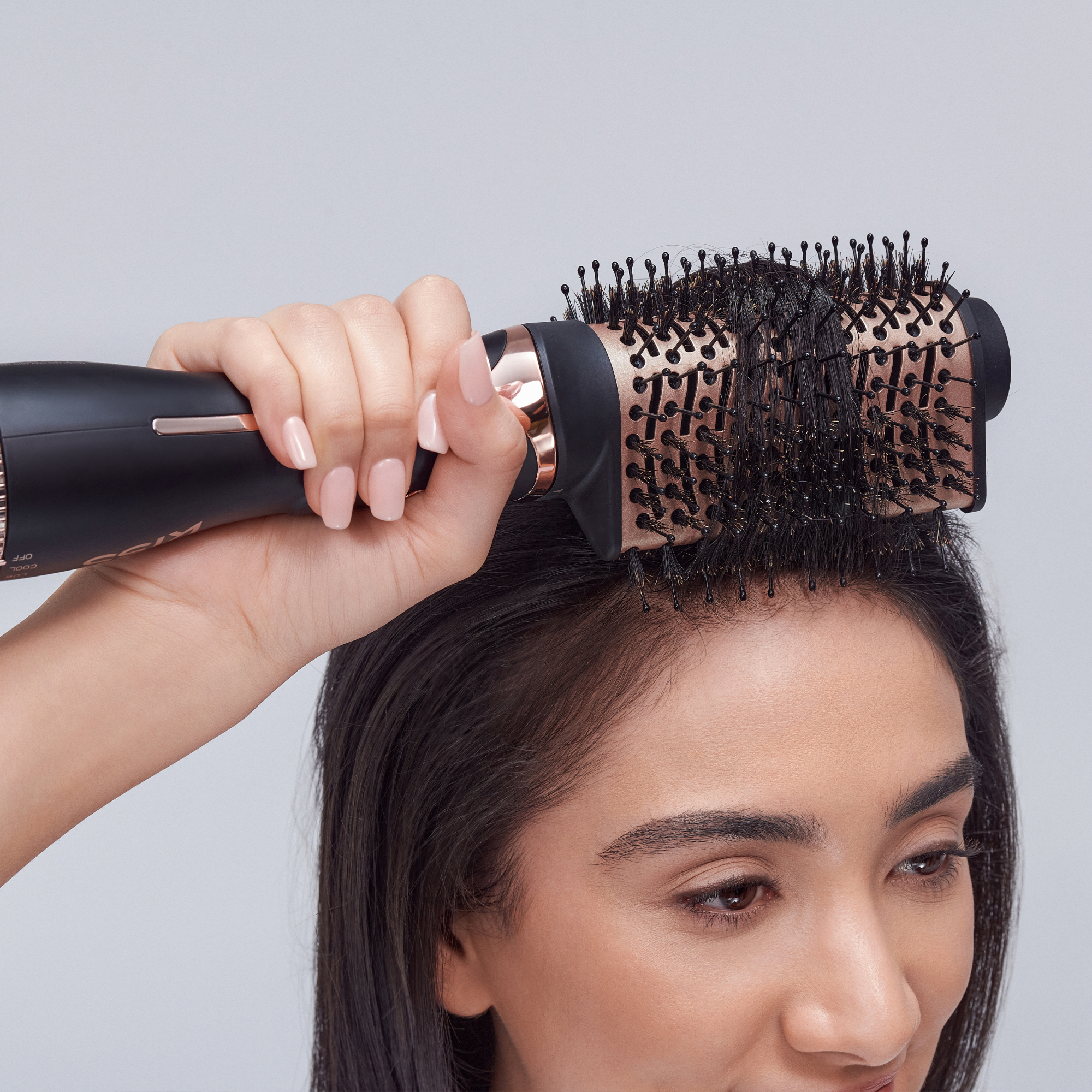 The image features a close-up of a person styling their hair using a round brush. The brush has a black handle and a barrel with bristles, and it appears to be designed for smoothing or volumizing hair. The individual has long, dark hair and is holding the brush close to their scalp, suggesting they are in the process of blow-drying or styling their hair. The background is a soft, neutral color, which keeps the focus on the hair and the styling tool.