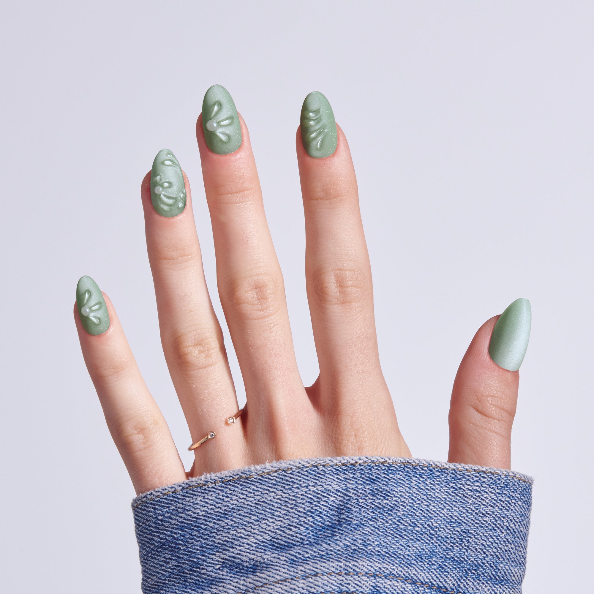 Sage green almond nails with white leaf designs on hand wearing denim sleeve.