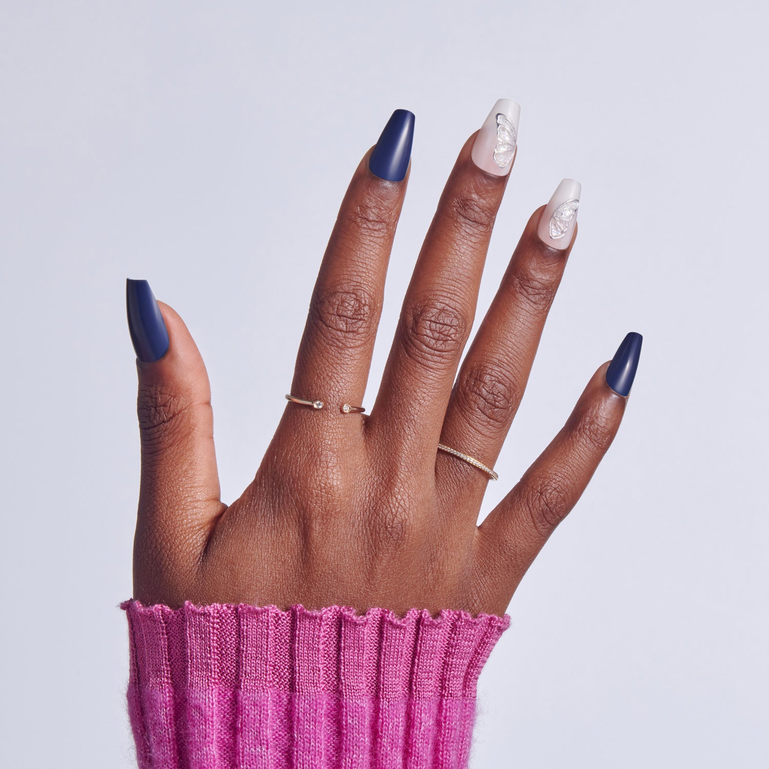 Hand with navy and white nail polish, wearing delicate rings, against pink sleeve.
